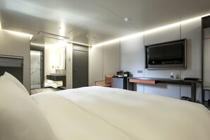 Una habitación de hotel moderna con una cama grande, un escritorio con televisor de pantalla plana, una pequeña zona de estar y un baño visible a través de una puerta abierta.