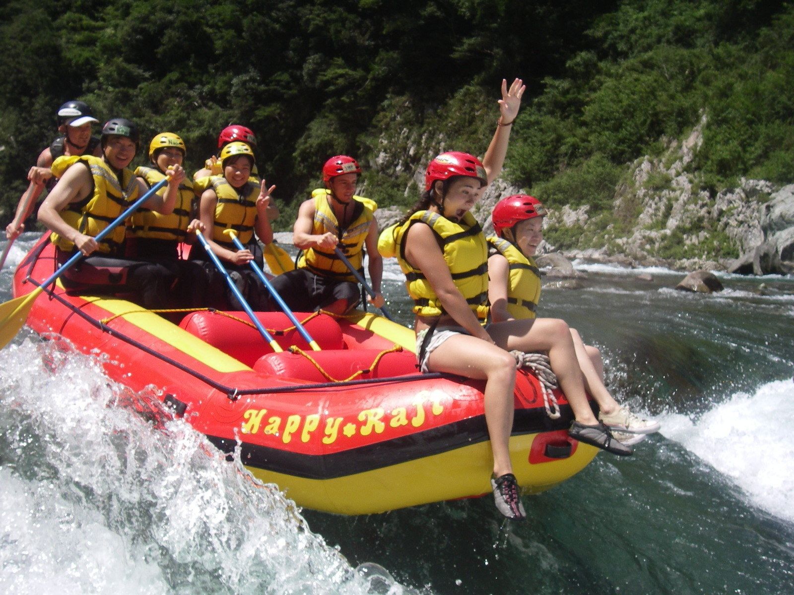 A group of people in a red and yellow raft with 'Happy Raft' written on it, navigating a river surrounded by greenery.