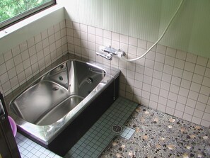 A bathroom with a large, rectangular, metallic bathtub, a showerhead, and a tiled floor with a mix of small and large tiles. - Happy Guest House (Otoyo)