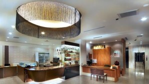 Un lobby moderno de hotel con un mostrador de recepción central, una amplia área de recepción de madera y una cocina visible al fondo.