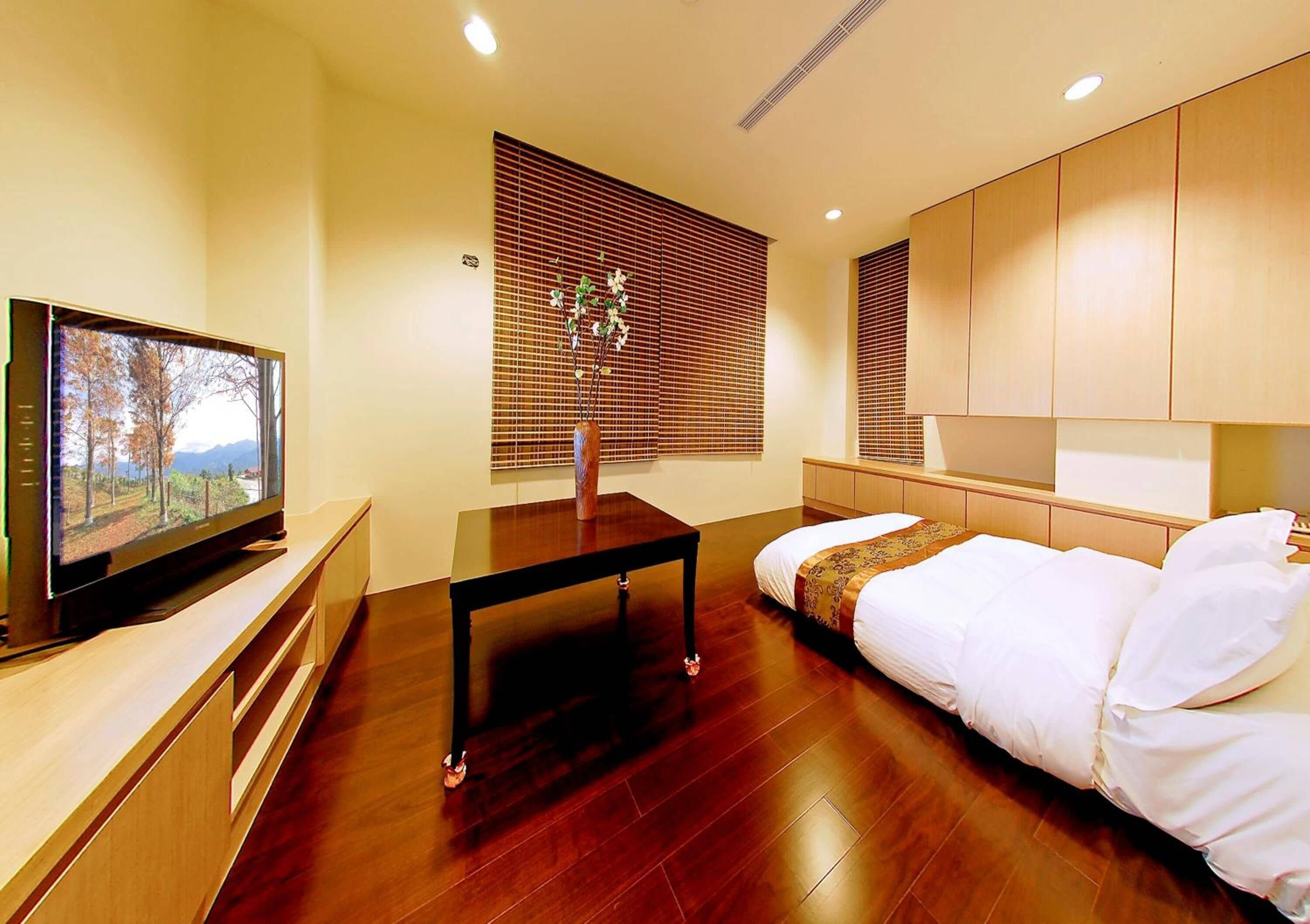 A modern hotel room with a wooden floor, a bed with white bedding, a TV on a wooden console, a small table, and a window with blinds.