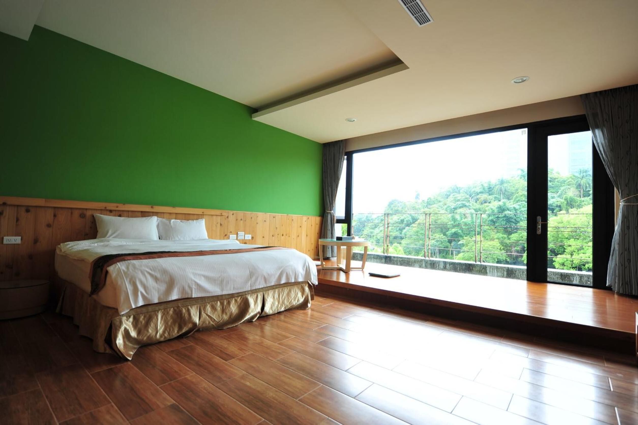 A bedroom with a wooden bed, green walls, and a large window offering a view of greenery.