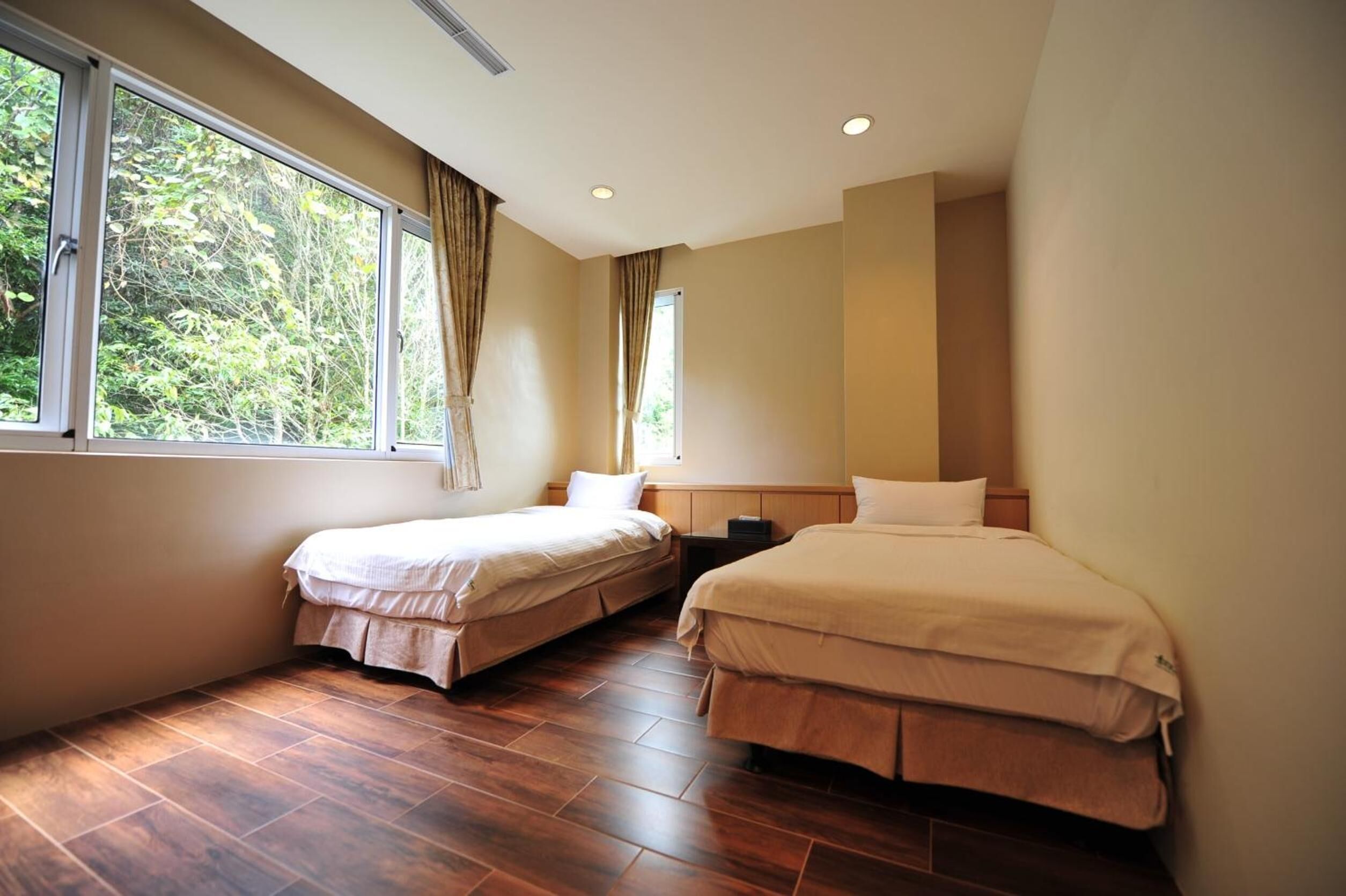 A hotel room with two beds, a large window with curtains, wooden flooring, and a neutral color scheme.