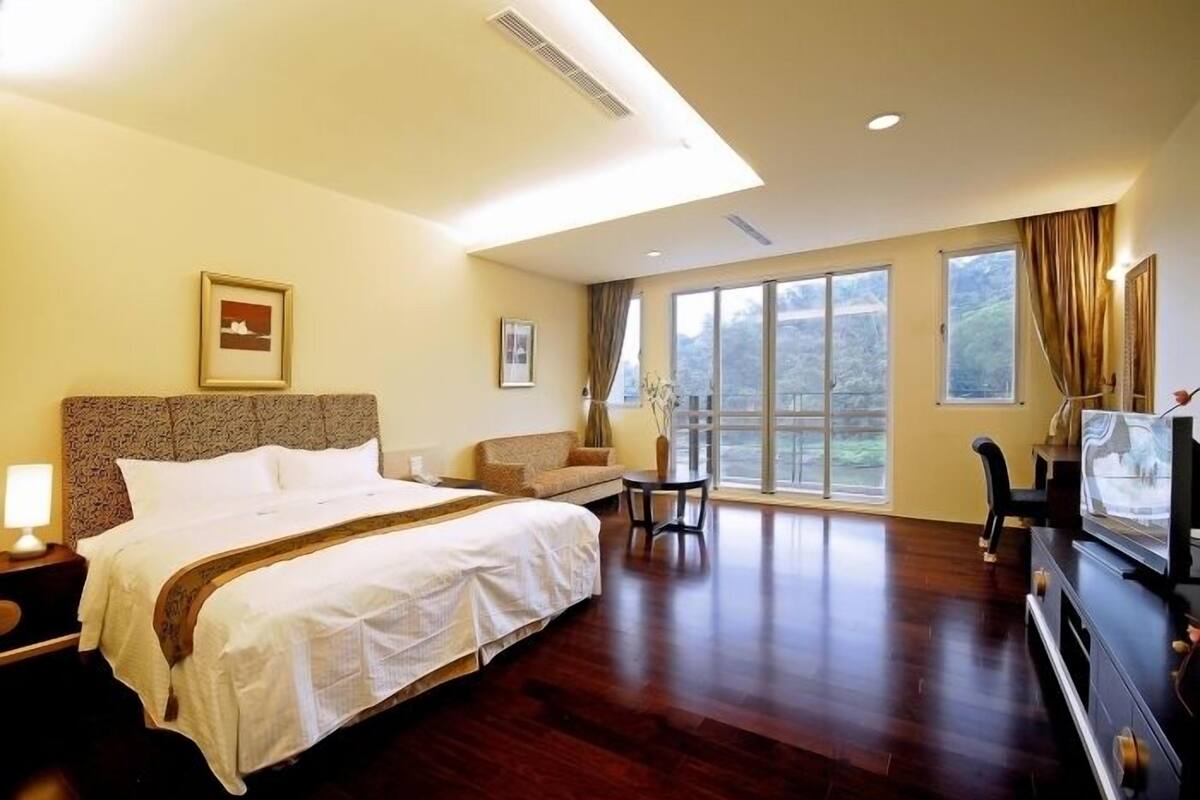 Une chambre d’hôtel avec un grand lit, un canapé, une petite table, une télévision et de grandes fenêtres donnant sur l’extérieur.