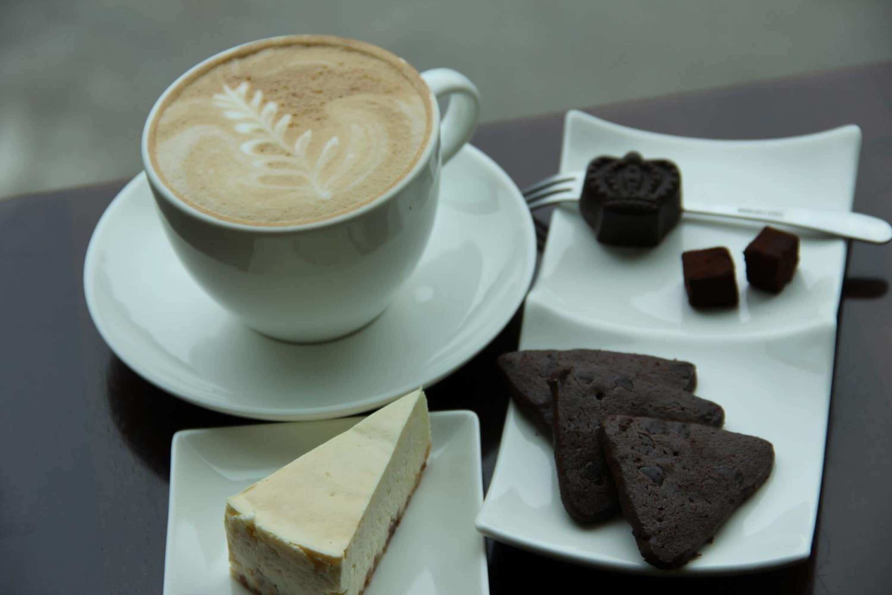 Una taza de café con decoración de latte, una porción de pastel de queso y galletas de chocolate sobre un plato blanco.