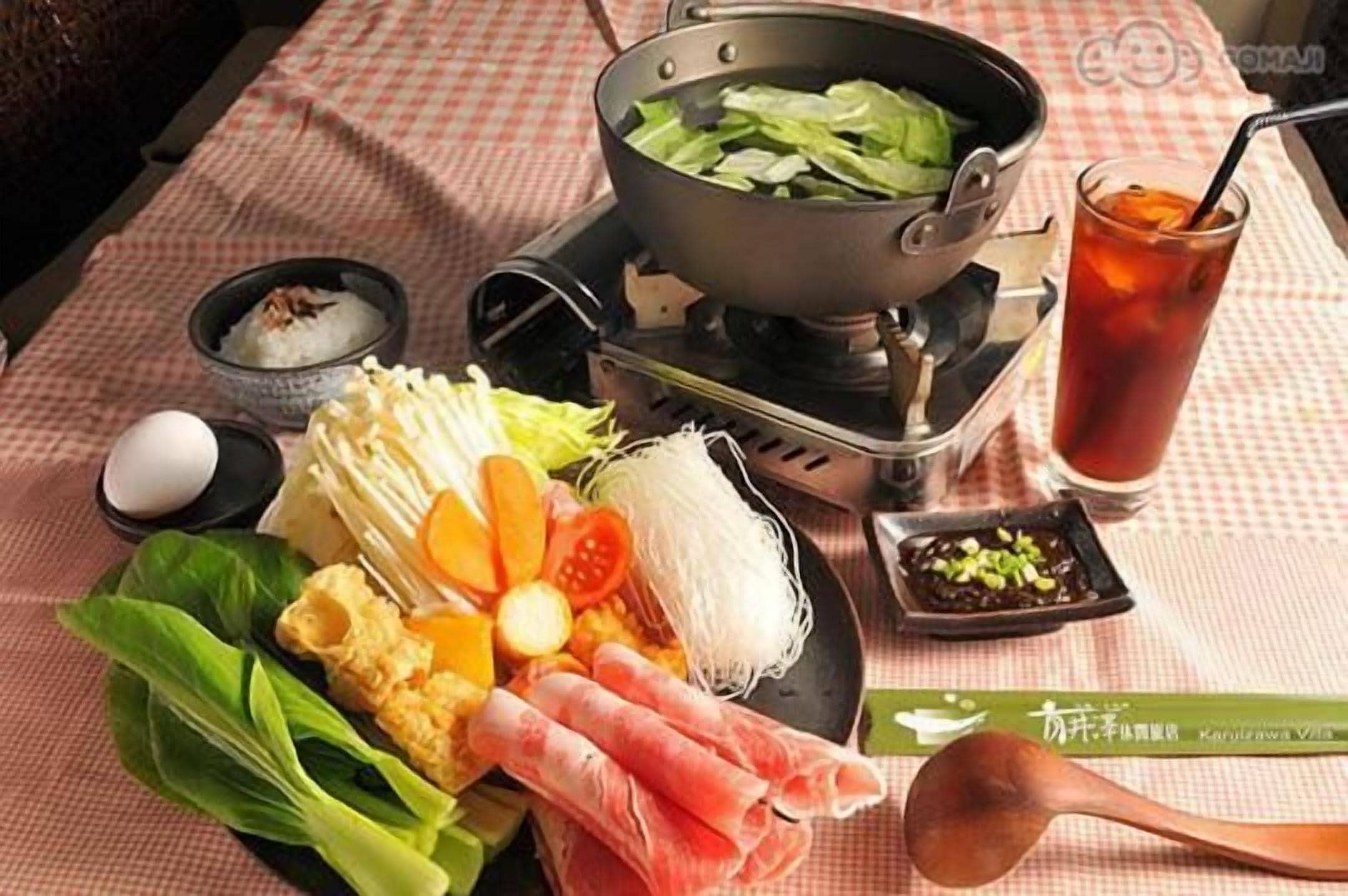 A table with a hot pot setup, various vegetables, a bowl of rice, a glass of iced tea, and condiments.