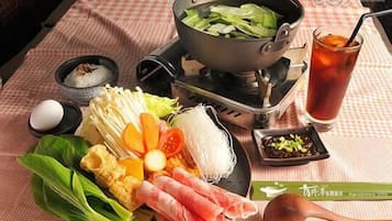 A table with a hot pot setup, various vegetables, a bowl of rice, a glass of iced tea, and condiments.