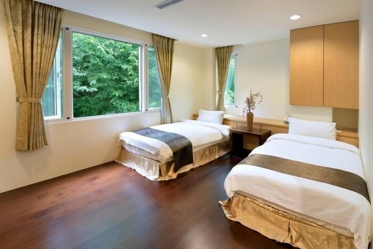 Une chambre d’hôtel avec deux lits, un plancher en bois, une fenêtre avec des rideaux, une table de chevet et une armoire.