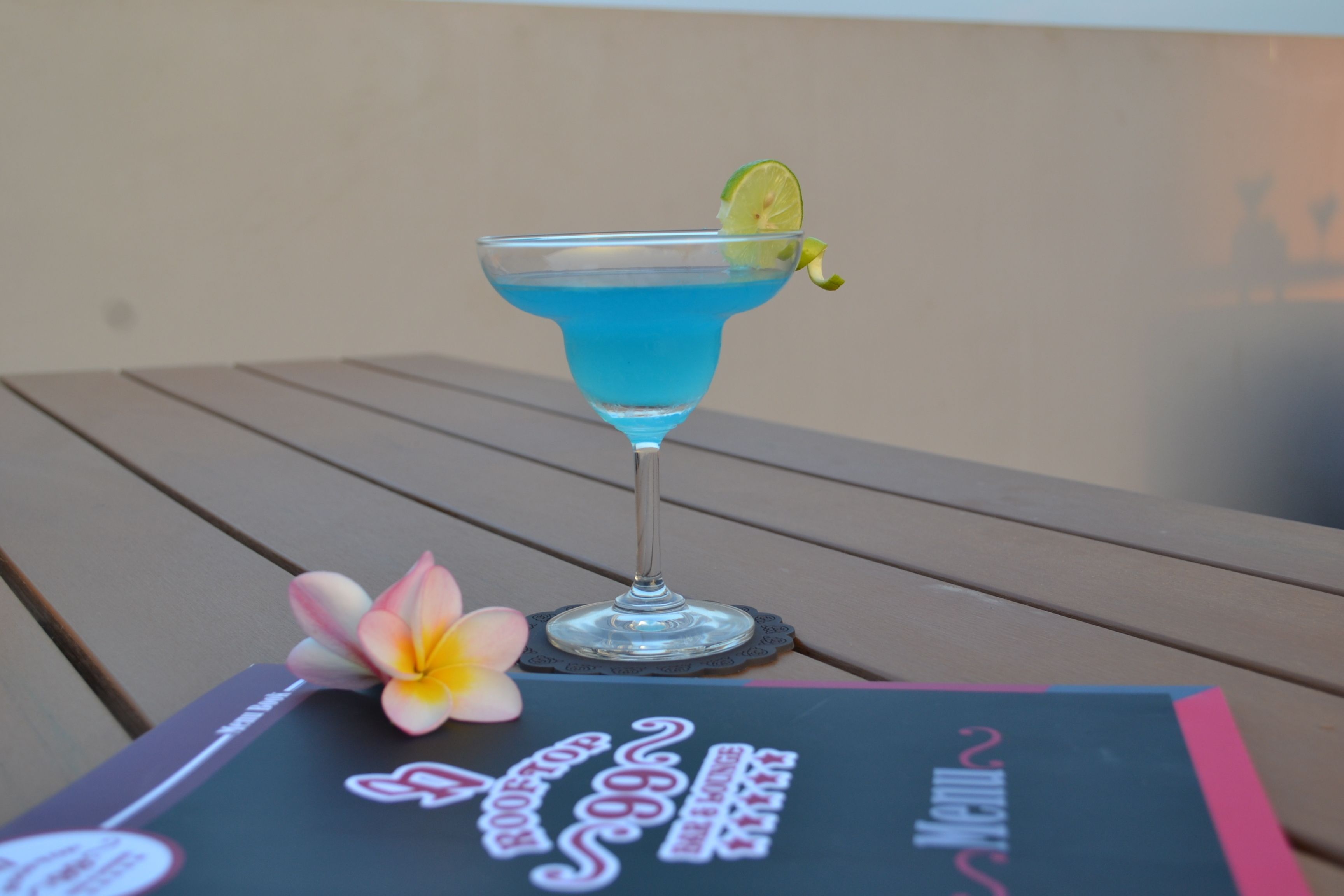 Um copo de cocktail com uma bebida azul, uma fatia de limão e uma flor sobre uma mesa.