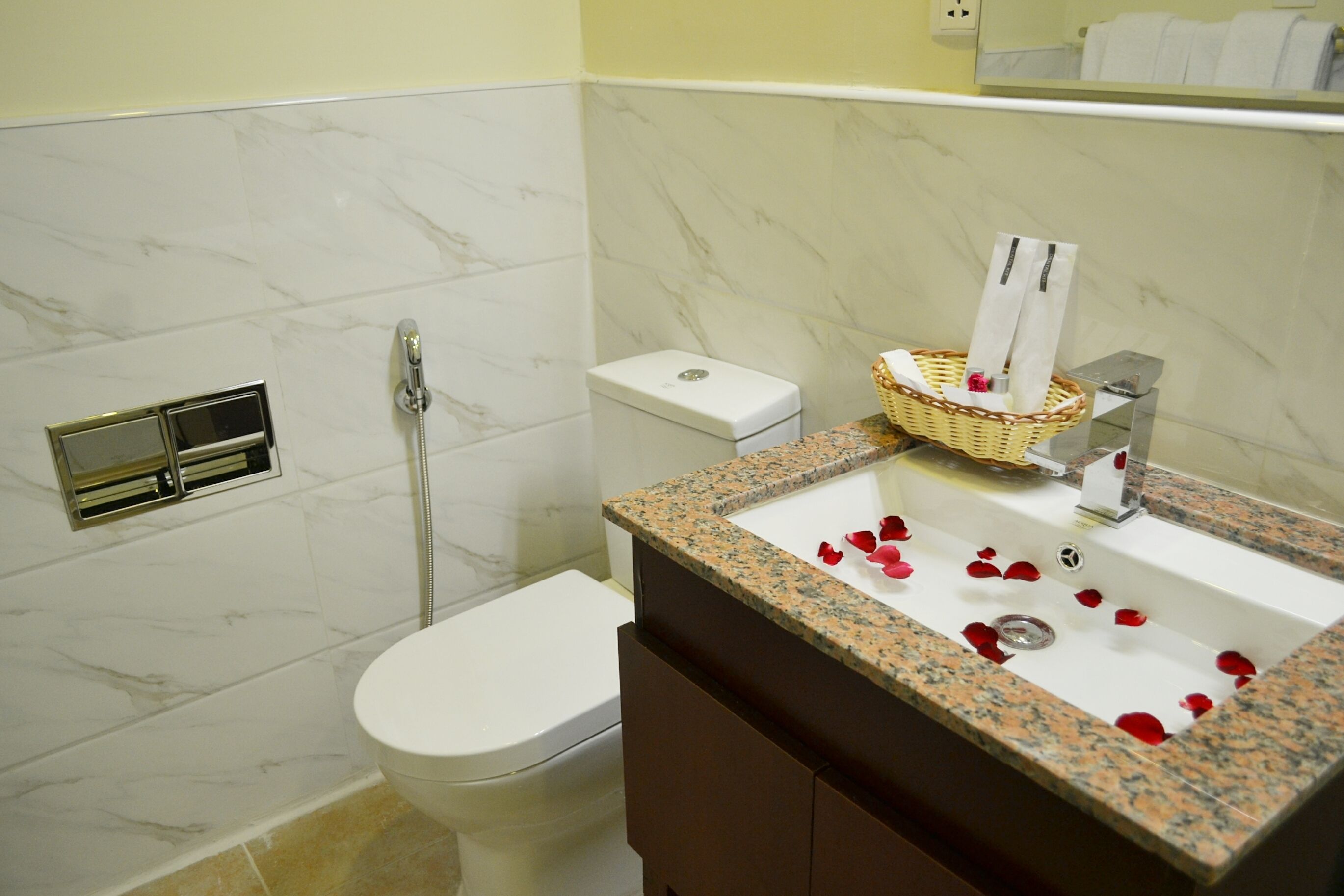 Un baño con un inodoro blanco, una pared de mármol, un lavabo con encimera de granito y una cesta sobre el lavabo.