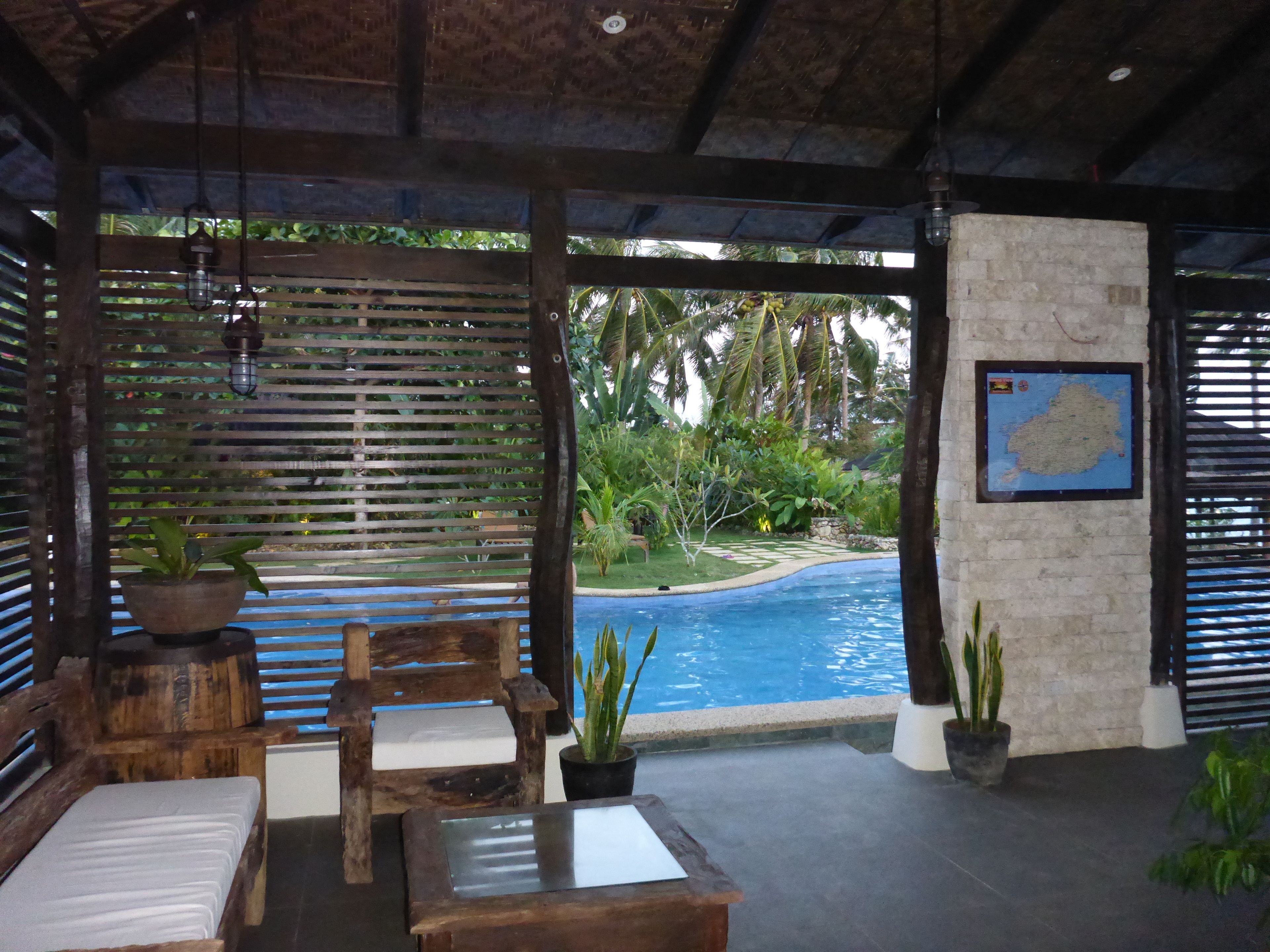 A poolside area with wooden furniture, a stone wall with a map, and a view of a tropical garden.