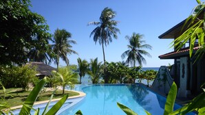 A clear blue swimming pool surrounded by lush greenery and palm trees, with a view of the ocean and a building with a thatched roof in the background.