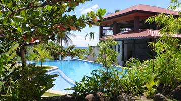 A swimming pool surrounded by tropical vegetation, a building with a red roof, and a view of the ocean.