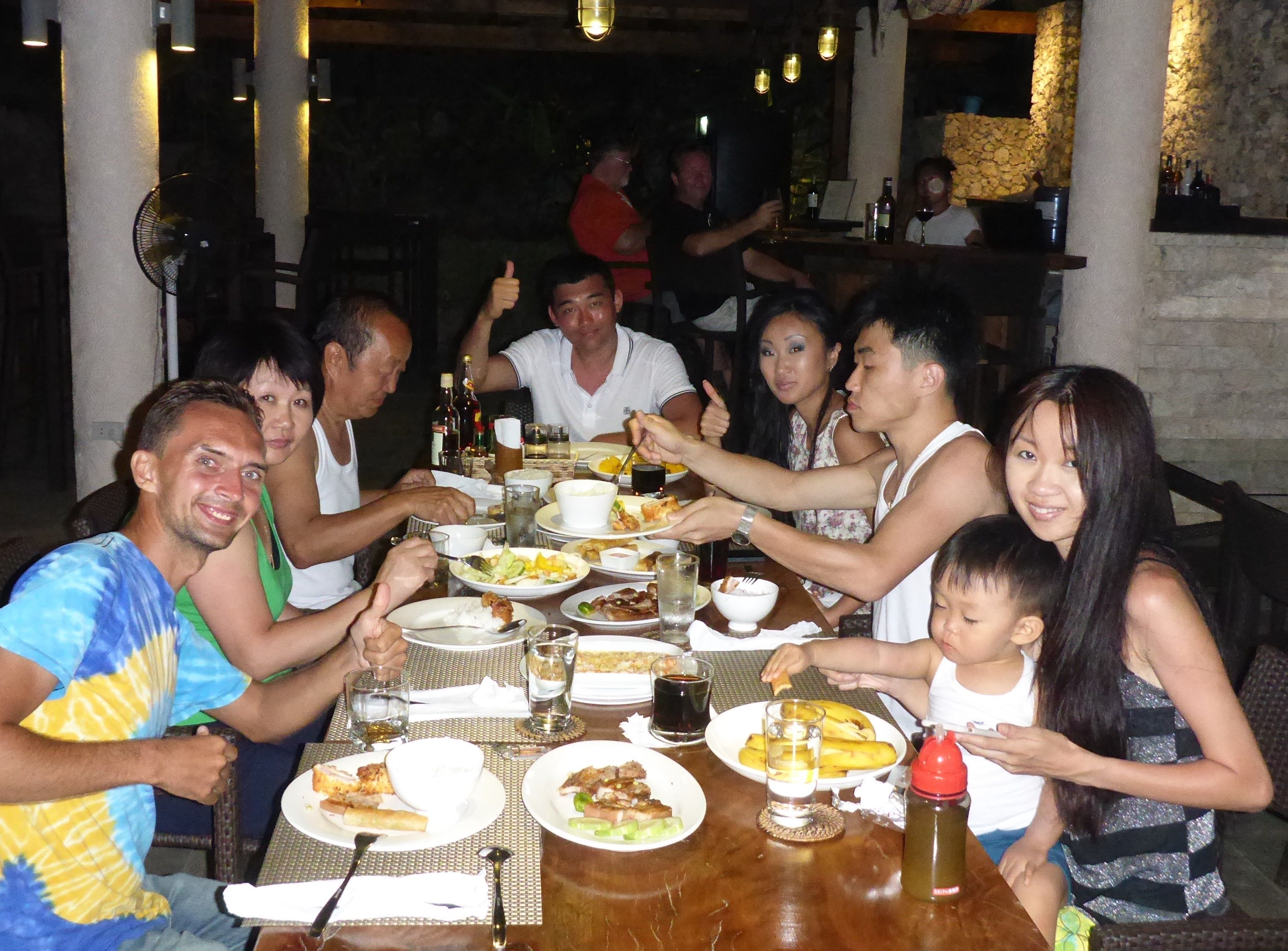 A group of people dining at an outdoor restaurant with a stone wall backdrop and hanging lights.