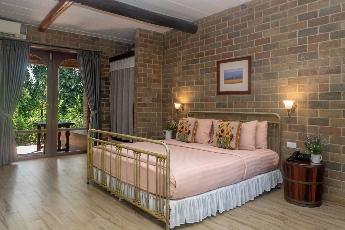 A bedroom with a brick wall, a bed with floral pillows, a nightstand with a lamp, and a view of greenery outside.