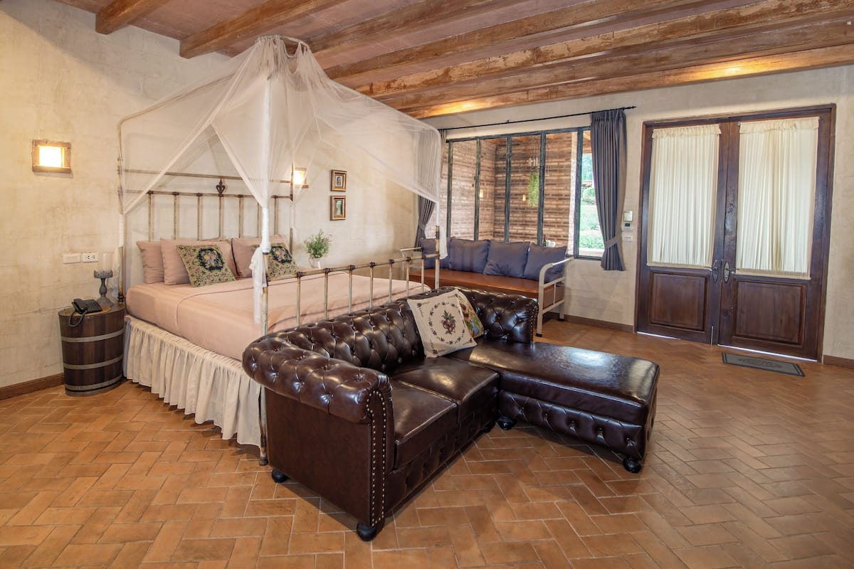 A bedroom with a four-poster canopy bed, a leather sofa, and a view of the outdoors through large windows.
