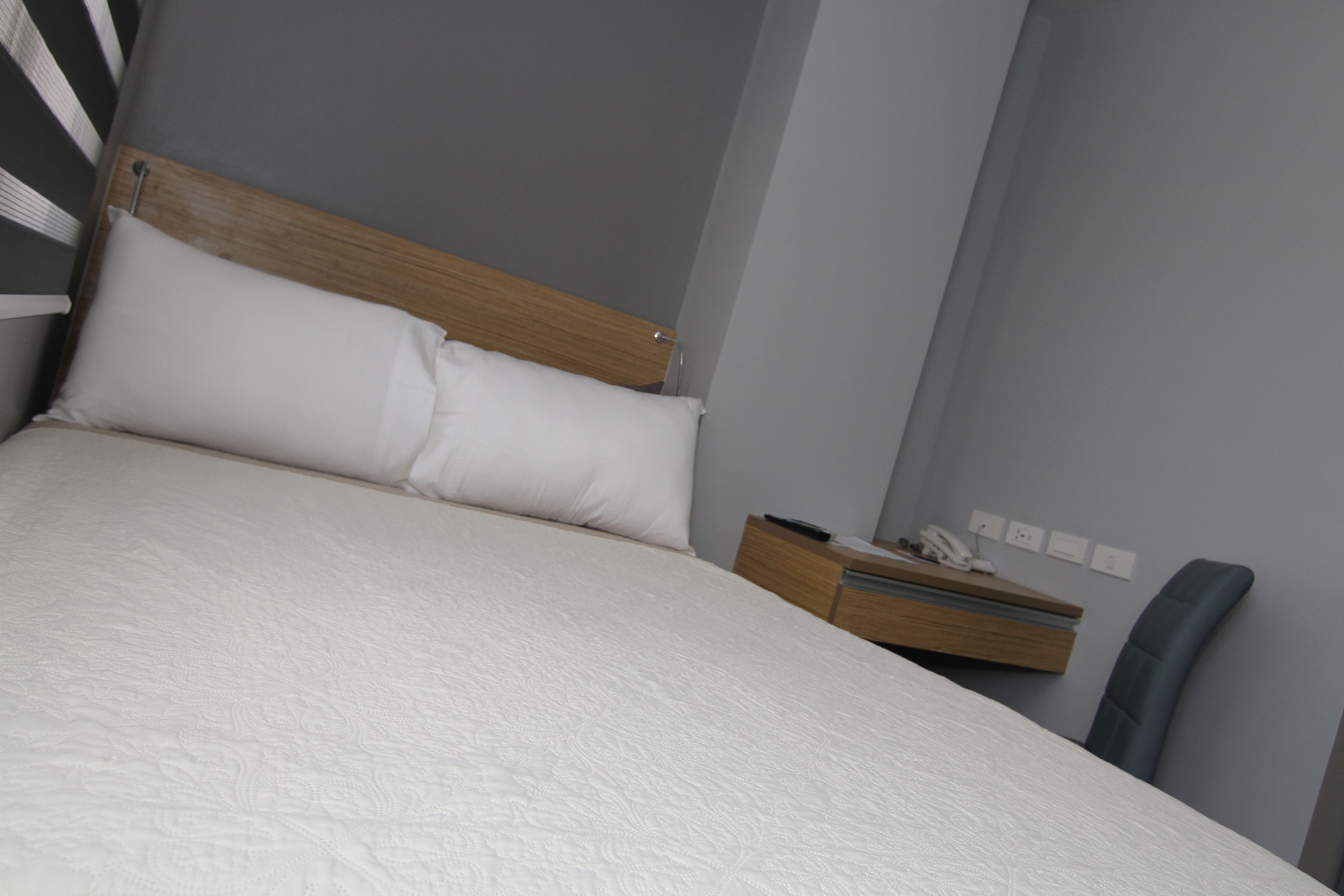 Una cama bien hecha con cabecera de madera y ropa de cama blanca en una habitación con pared gris y una pequeña mesita de noche con un teléfono y un control remoto.