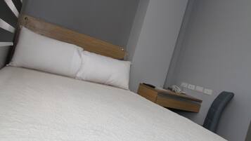 Una cama bien hecha con cabecera de madera y ropa de cama blanca en una habitación con pared gris y una pequeña mesita de noche con un teléfono y un control remoto.