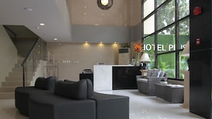Un hall d’hôtel moderne avec un canapé d’angle noir, un comptoir d’accueil et de grandes fenêtres donnant sur un paysage verdoyant.