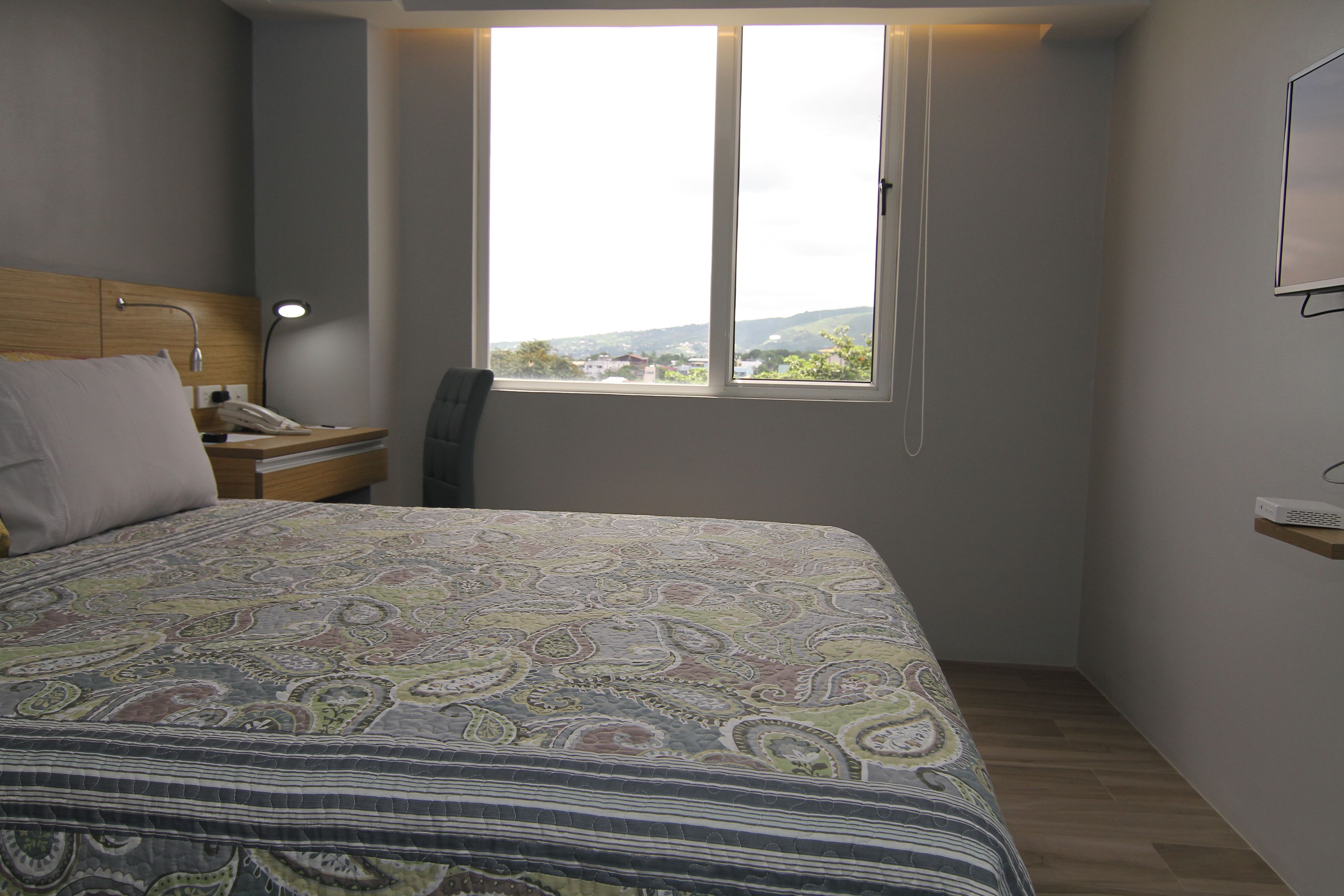 A bedroom with a bed, a nightstand, a desk, a chair, and a window with a view.