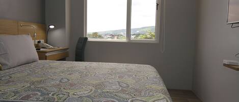 A bedroom with a bed, a nightstand, a desk, a chair, and a window with a view.