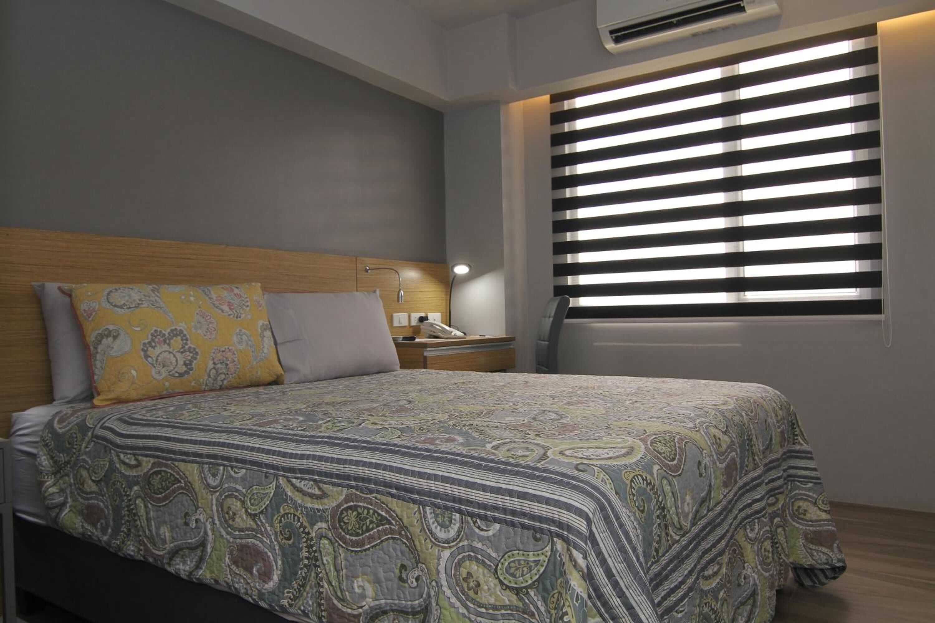 Quarto com cama, mesa de cabeceira, luminária e janela com persianas.