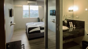 Une chambre d’hôtel moderne comprenant un lit, une grande fenêtre, un téléviseur à écran plat, une salle de bain avec un miroir et un lavabo, ainsi qu’une petite table avec une chaise.