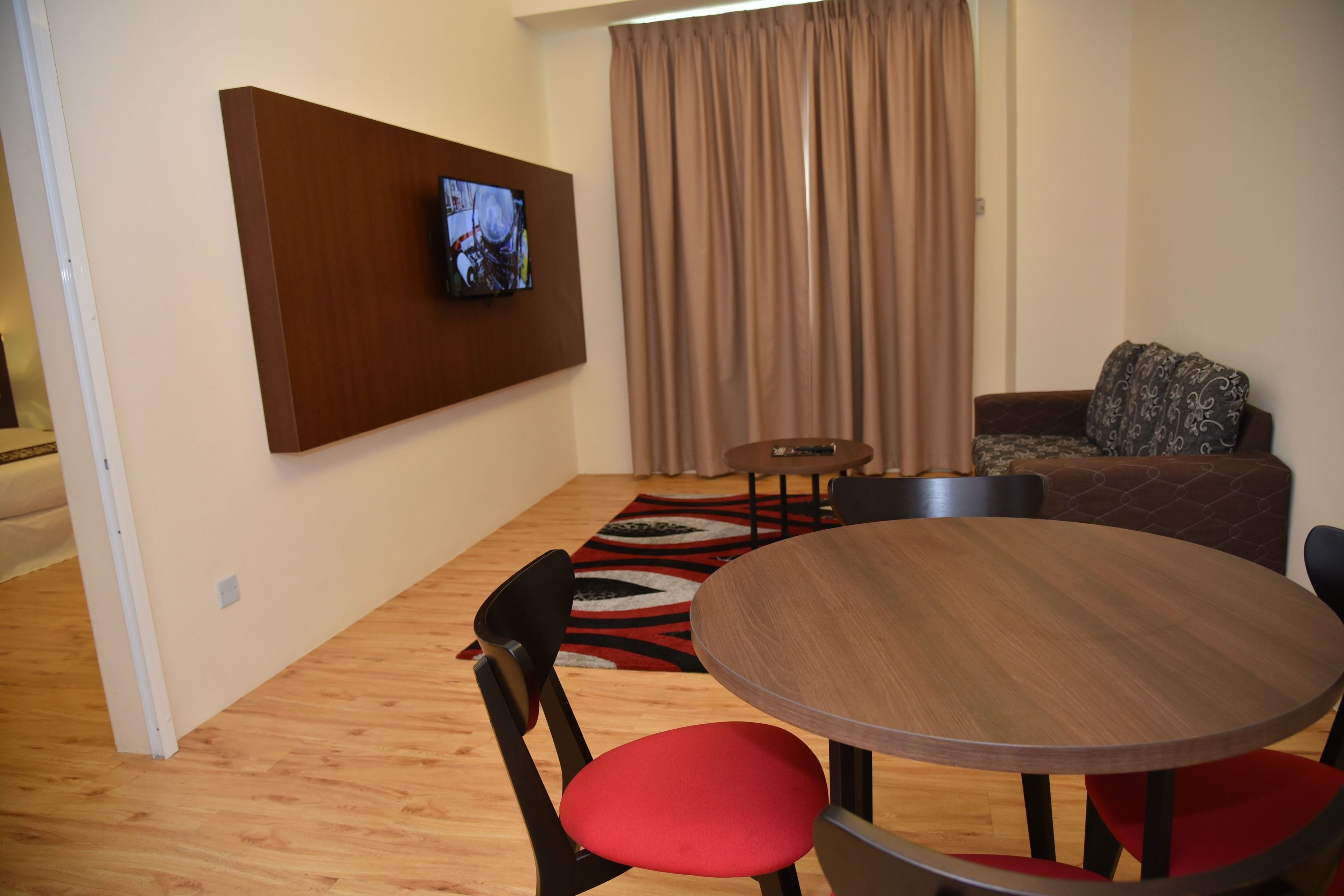 A modern living room with a round wooden table, red cushioned chairs, a television mounted on a wooden panel, and a patterned rug.