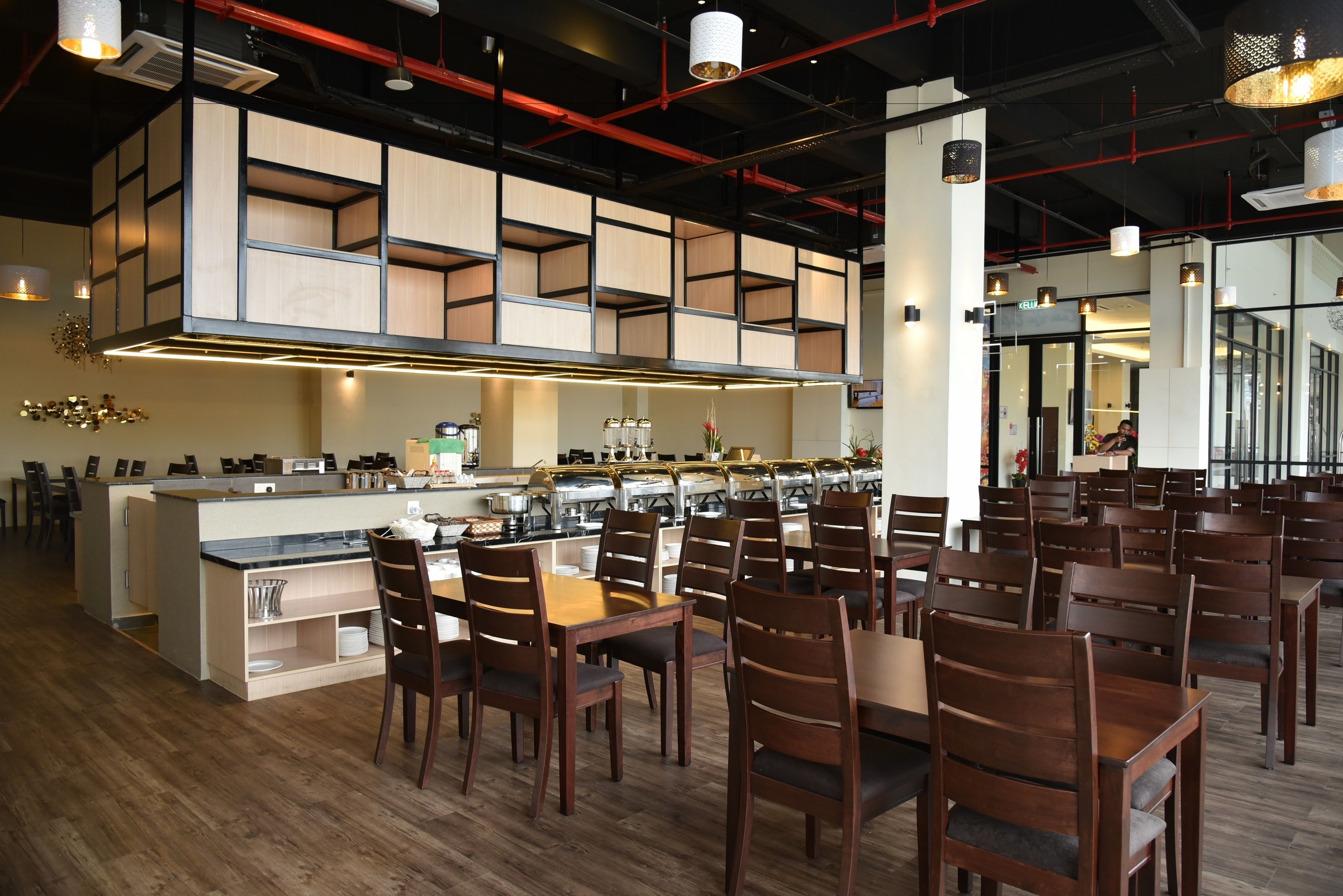 A modern restaurant interior with wooden flooring, a central island with kitchen appliances, and multiple dining tables with chairs.
