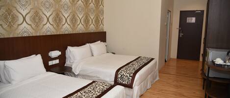 Quarto de hotel com duas camas, cabeceira de madeira, papel de parede estampado, piso de madeira, uma porta e uma pequena mesa com abajur.
