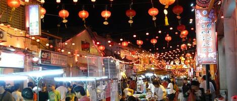 Un mercado callejero bullicioso de noche, con faroles rojos colgando y varios puestos que venden productos.