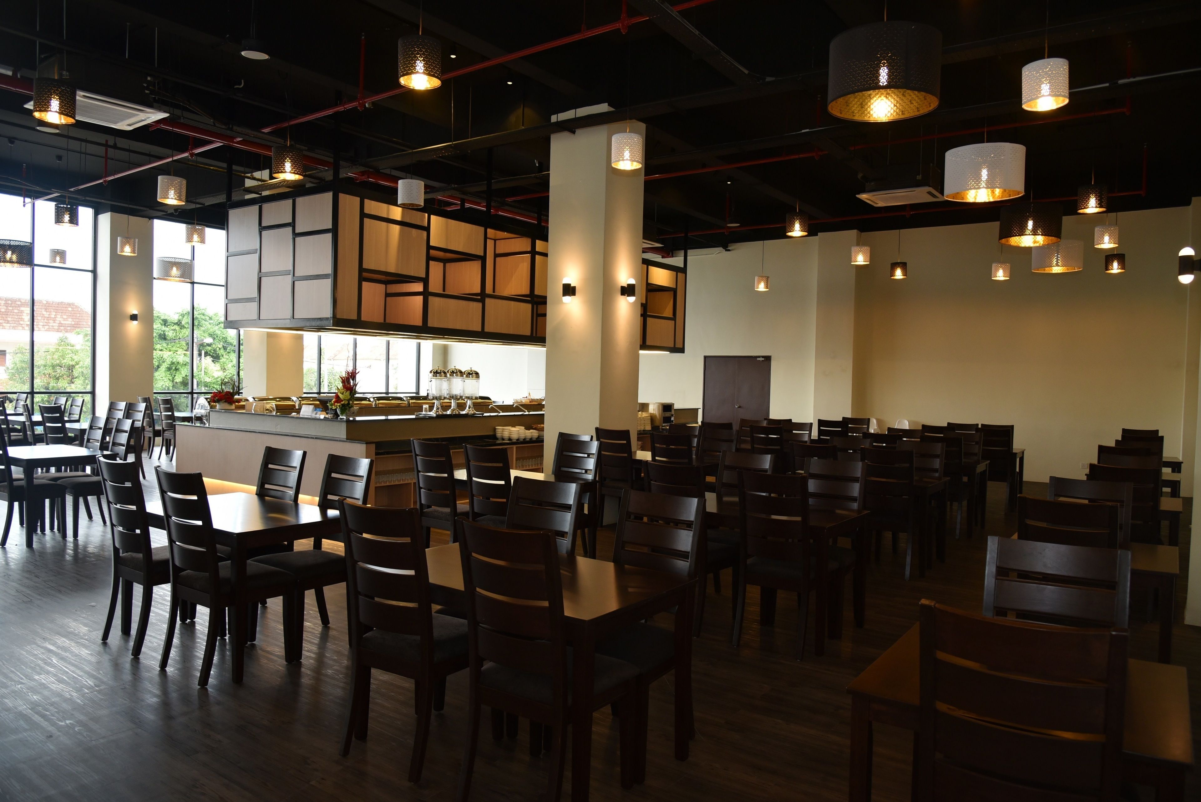 Un restaurant spacieux, doté de nombreuses tables et chaises, d’un espace cuisine et d’un éclairage suspendu.