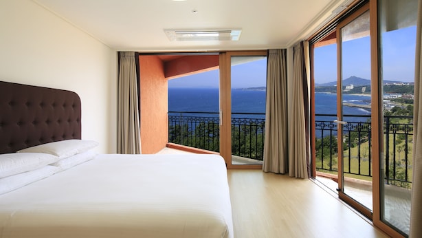 A hotel room with a large bed, a view of the sea, and a balcony with a railing. - Jeju Booyoung Resort (Seogwipo)