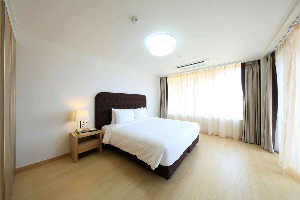 Une chambre d’hôtel bien rangée, avec un grand lit, une table de chevet avec une lampe et une fenêtre avec des rideaux.