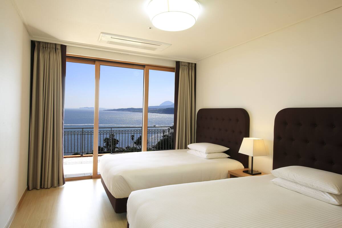 Une chambre d’hôtel avec deux lits, une grande fenêtre donnant sur la mer et un luminaire au plafond.