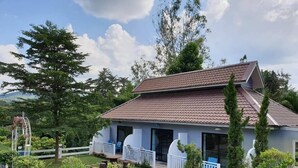 Une maison à deux étages, avec un toit recouvert de tuiles, entourée d’un jardin où poussent différentes plantes et arbres.