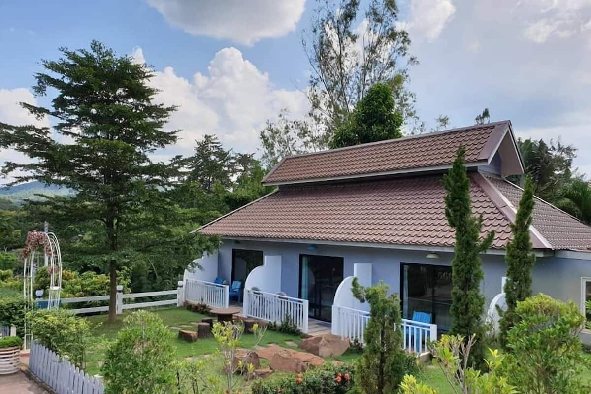 Una casa de dos pisos con techo de tejas, rodeada por un jardín con diversas plantas y árboles.