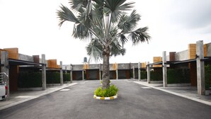 Um estacionamento com uma palmeira central, rodeado por vegetação, pilares de concreto e um carro branco estacionado à esquerda.