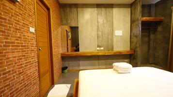 A hotel room with a brick wall, a wooden door, a large mirror, a bed with white linens, and a small trash bin.
