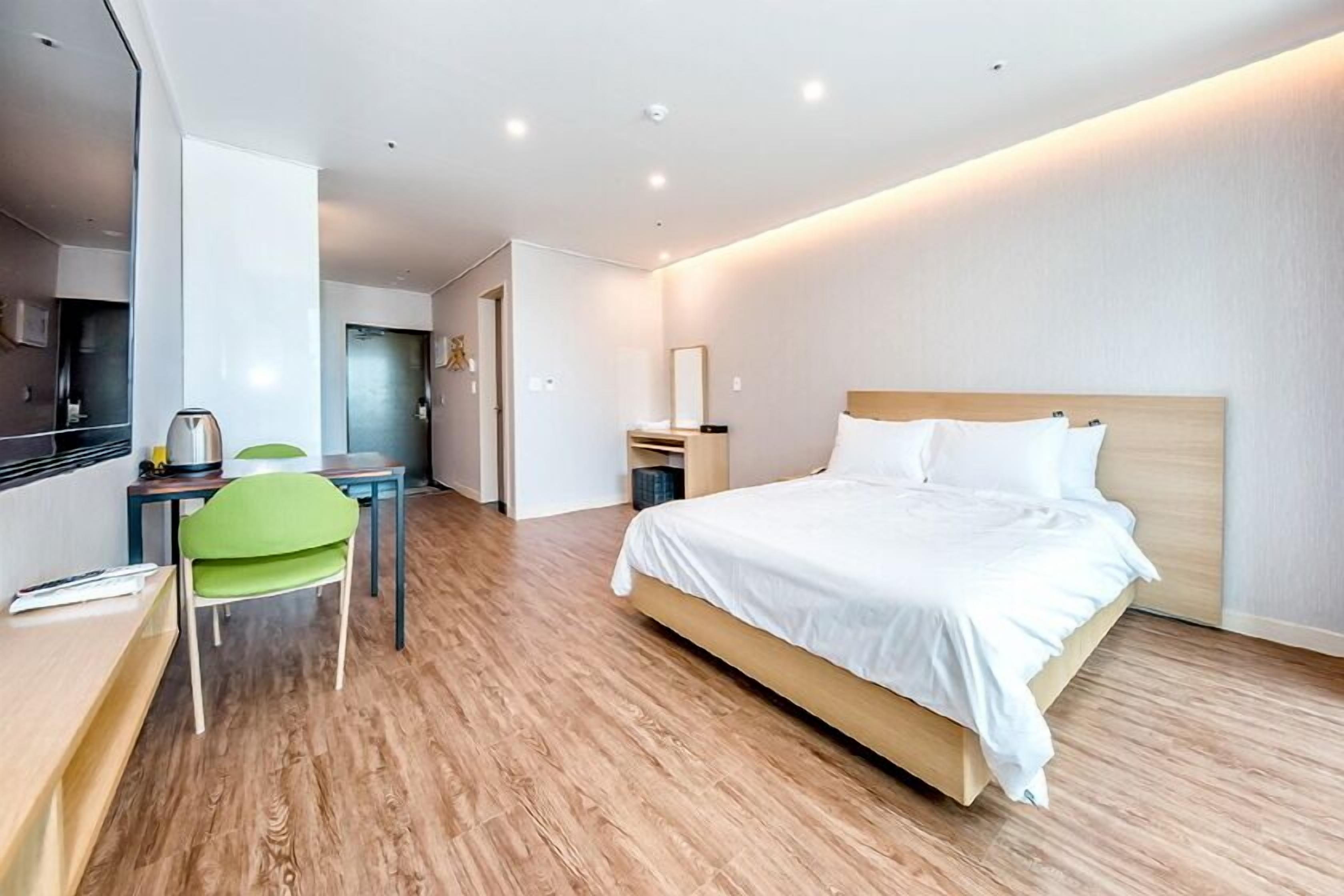 A modern hotel room with a wooden floor, a bed with white linens, a desk with a chair, and a small table with a kettle.