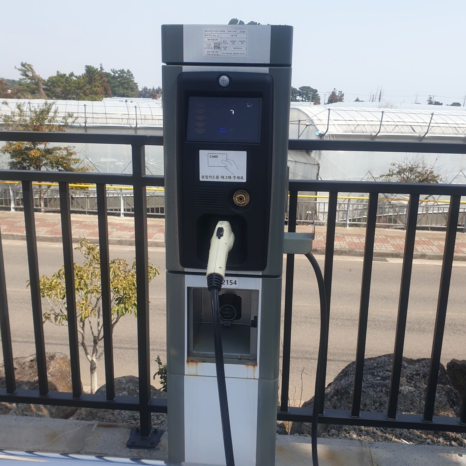 Estación de carga pública para vehículos eléctricos con pantalla digital, lector de tarjetas y un cable conectado a un vehículo.