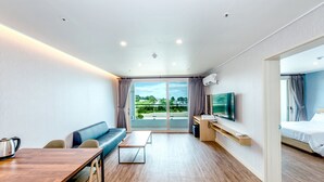 Een moderne hotelkamer met een houten vloer, een bank, een eettafel, een televisie en een balkon met uitzicht op groen.