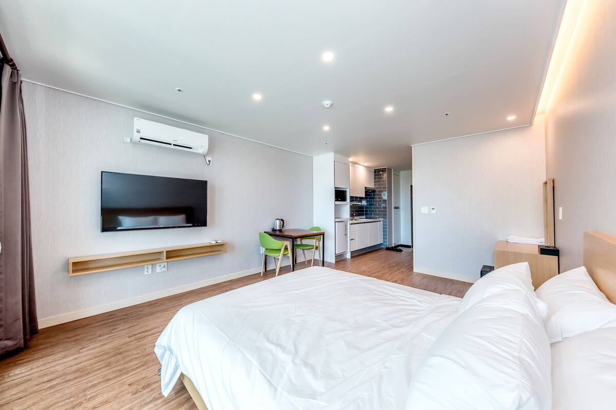 Ein modernes Hotelzimmer mit einem großen Bett, einem flachbildfernseher an der Wand, einer kleinen Küchenzeile und einer Essecke mit Tisch und Stühlen.