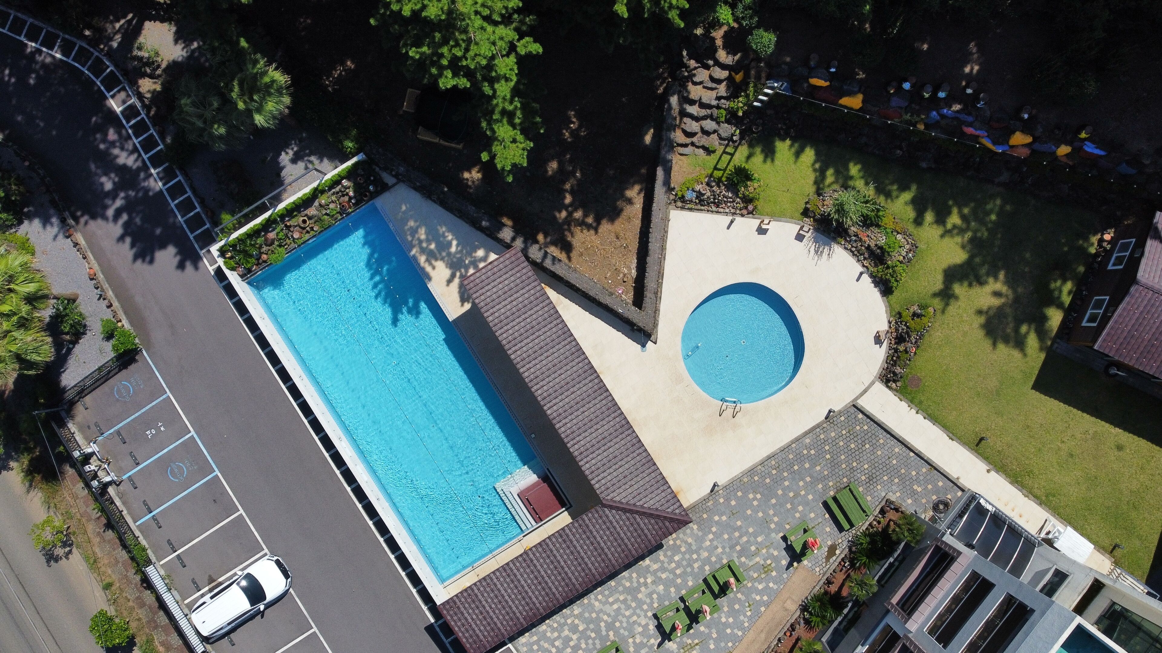 An aerial view of a swimming pool with a circular hot tub, surrounded by a paved deck and greenery.