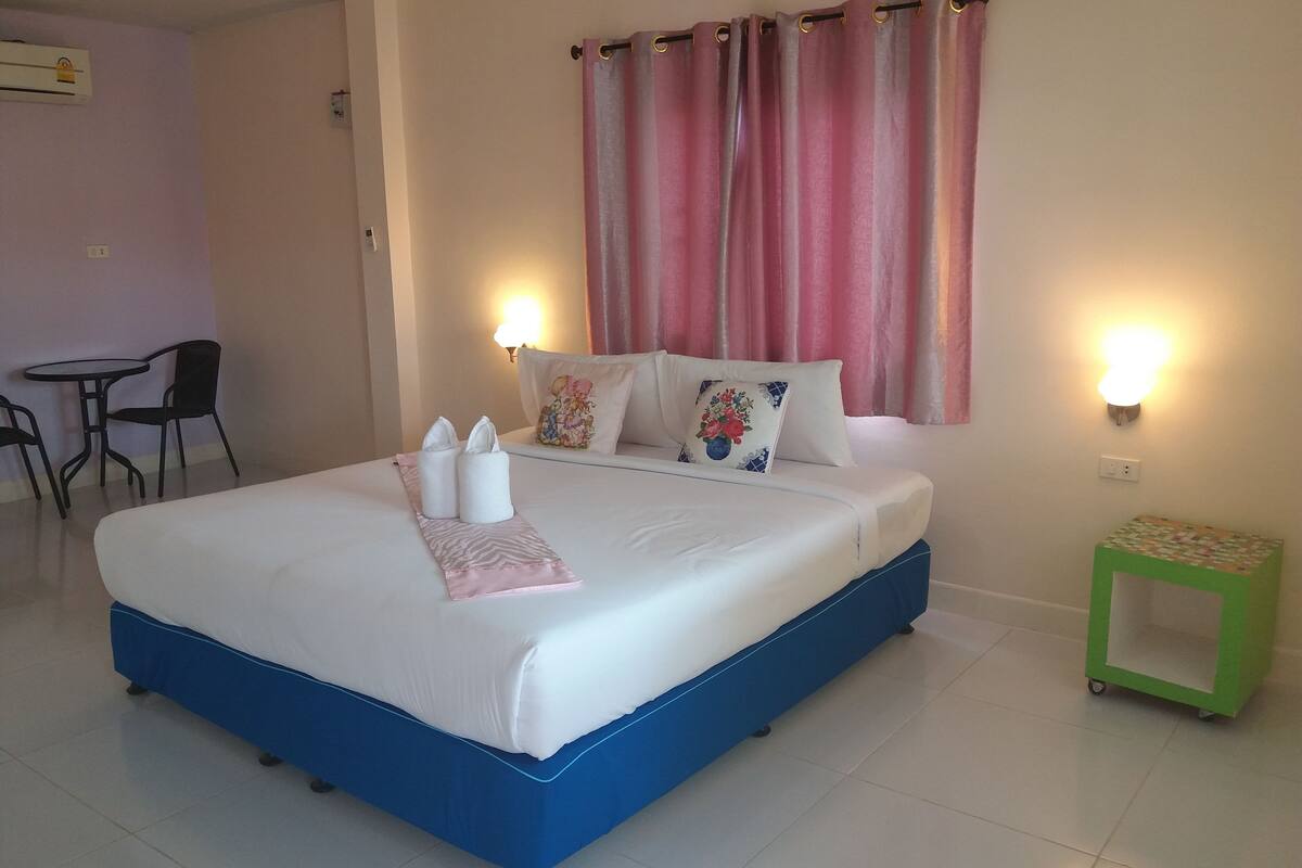 A neatly made bed with pillows and a pink towel, a small green side table, and a chair in a room with a tiled floor.