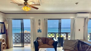 A spacious living room with a large window offering a view of the ocean. There is a ceiling fan, a wooden coffee table, and a sofa with cushions. - Araha Resort ARAPANA (Chatan)