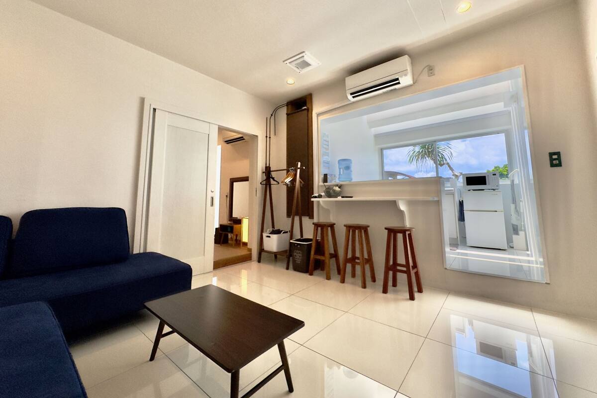 A modern living room with a sofa, a coffee table, and a kitchen area with stools and a refrigerator.
