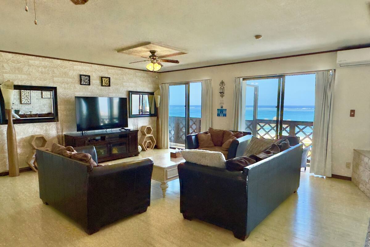 A spacious living room with a large flat-screen TV, comfortable seating, and a balcony with a view of the ocean.