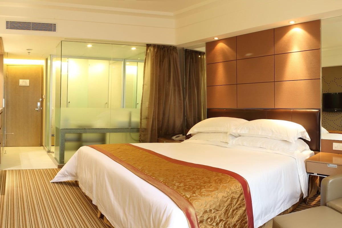 A hotel room with a bed, a desk, a chair, a TV, and a bathroom with a glass door.