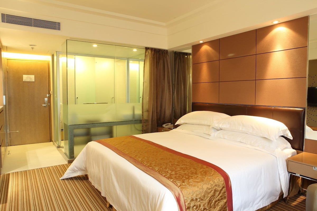 A hotel room with a large bed, a bedside table, a mirror, and a glass door leading to a shower area.